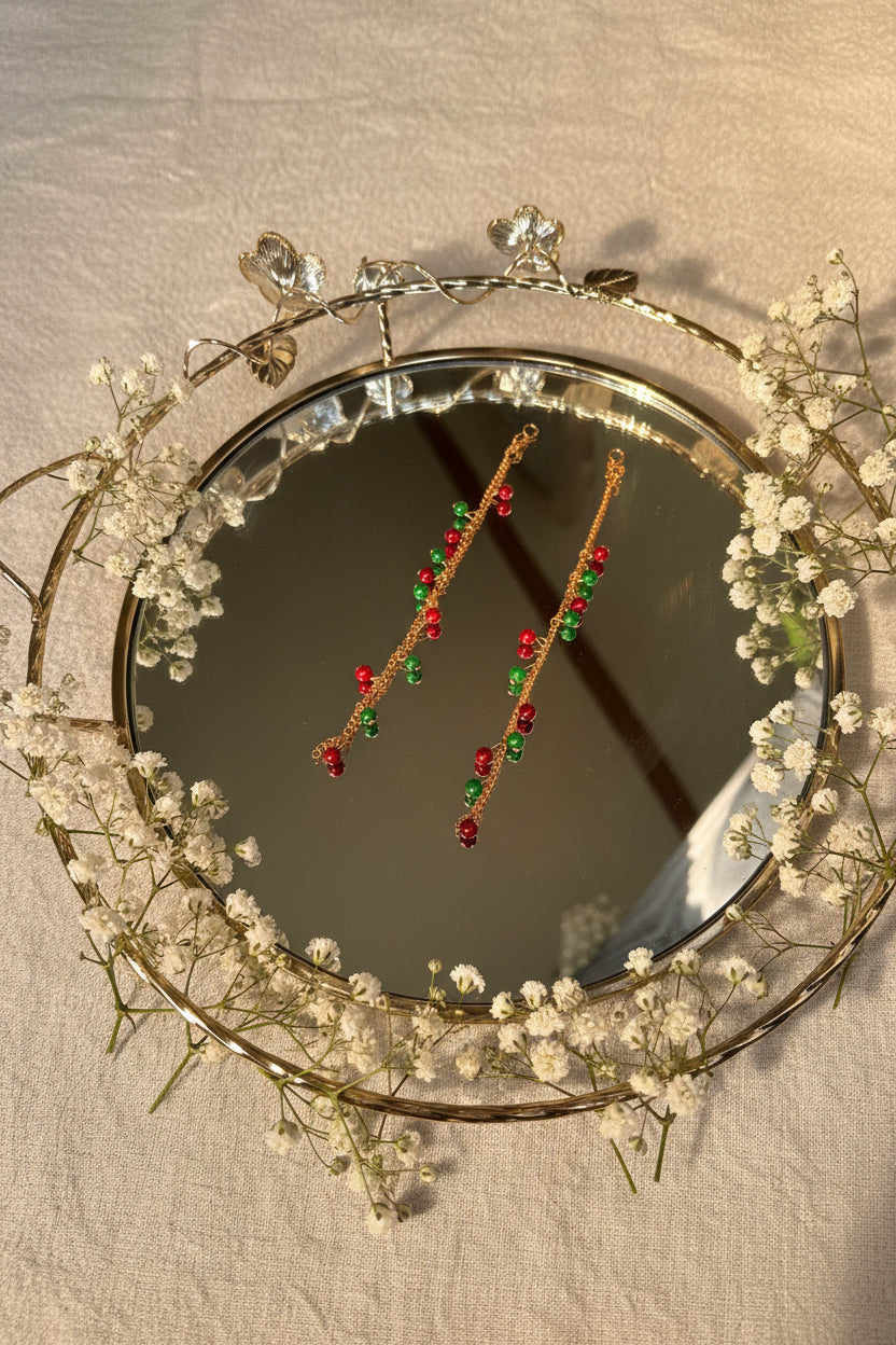 A pair of long gold-toned beaded jhumkha hair chains decorated with alternating red and green beads, displayed on a reflective silver tray.
