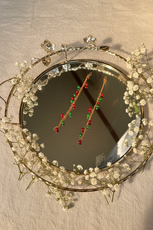 A pair of long gold-toned beaded jhumkha hair chains decorated with alternating red and green beads, displayed on a reflective silver tray.