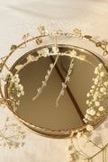 A close-up of a decorative oval mirror with a gold vine and leaf frame, reflecting two long, sparkling clear beaded strands or jhumkha chains against a light neutral background.