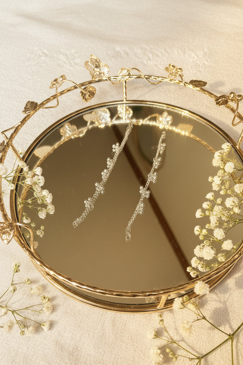 A close-up of a decorative oval mirror with a gold vine and leaf frame, reflecting two long, sparkling clear beaded strands or jhumkha chains against a light neutral background.