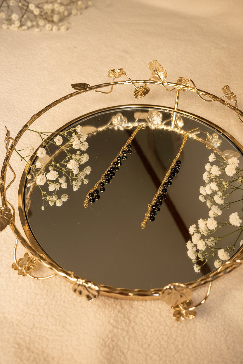 A pair of gold jhumkha hair chain earrings with black bead clusters, shown hanging in the reflection of a decorative round mirror with a gold floral frame.