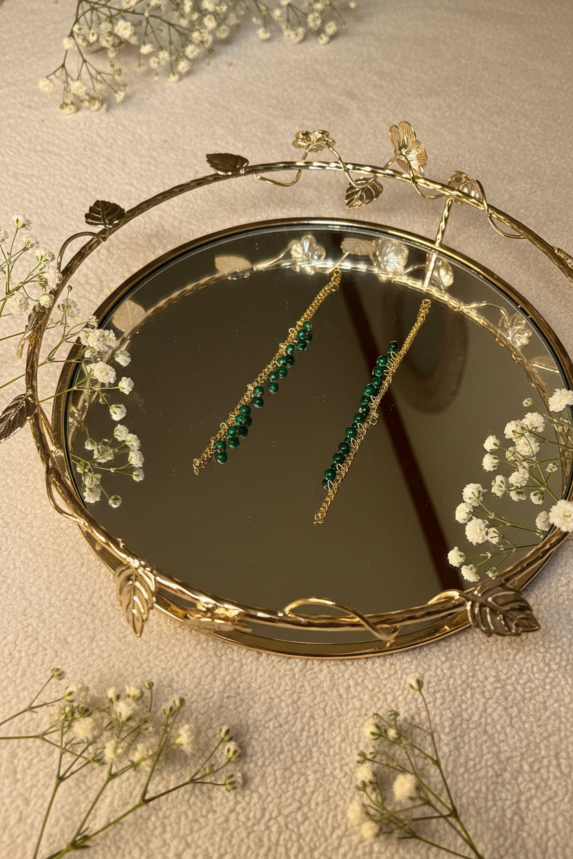 A pair of gold jhumkha hair chain dangle earrings with green gemstone beads, shown hanging in the reflection of a decorative gold-framed mirror.