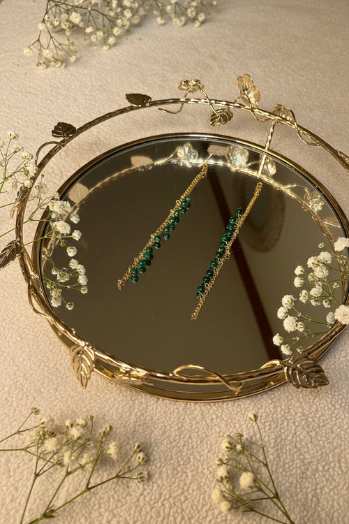 A pair of gold jhumkha hair chain dangle earrings with green gemstone beads, shown hanging in the reflection of a decorative gold-framed mirror.