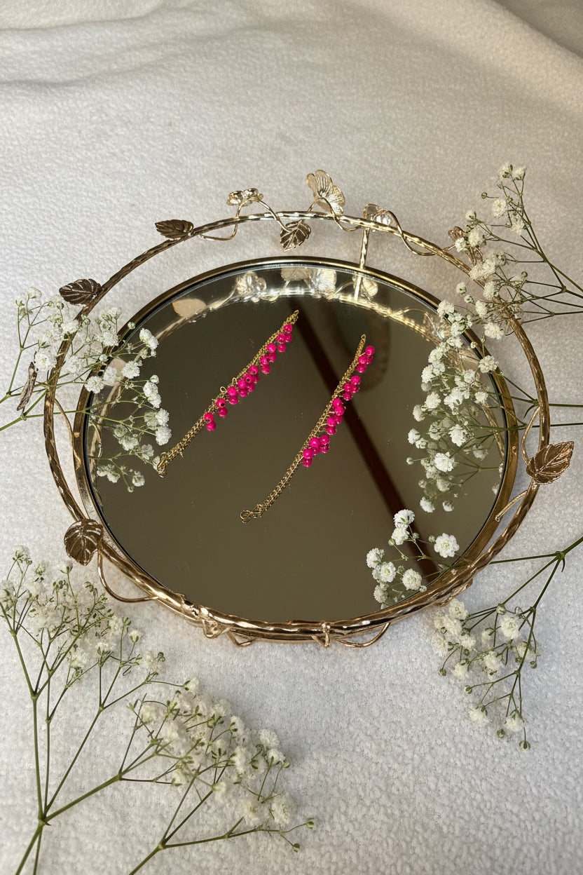 Gold jhumkha hair chain headband with bright pink flower embellishments, shown in a mirror reflection on a light textured surface.