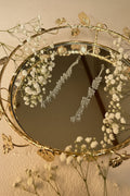 A pair of silver-toned statement jhumkha chain with clear rhinestone-like crystals, displayed on a round decorative gold mirror with a vine and leaf border.