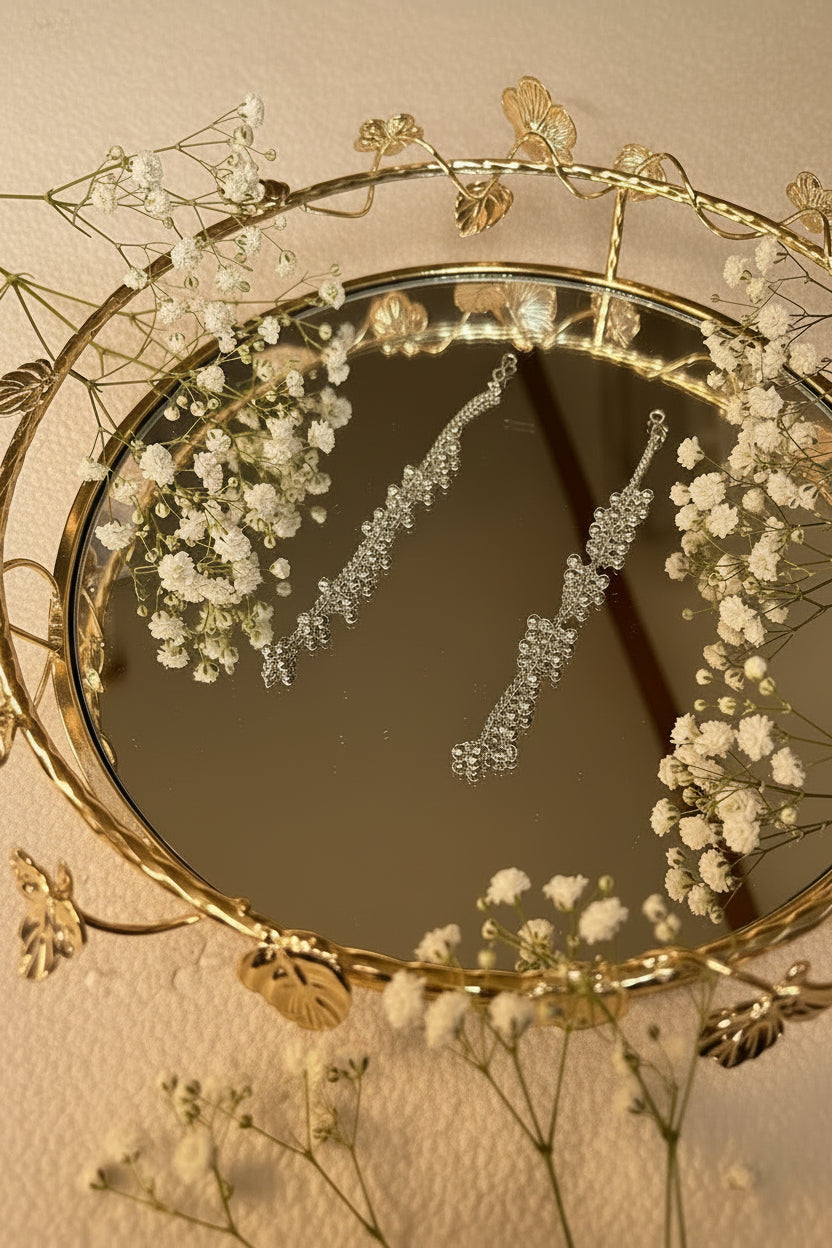A pair of silver-toned statement jhumkha chain with clear rhinestone-like crystals, displayed on a round decorative gold mirror with a vine and leaf border.