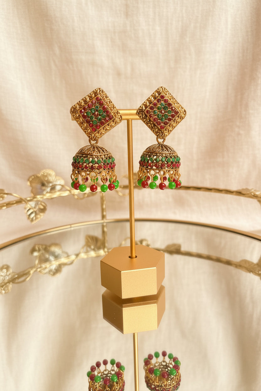 A pair of ornate gold-toned Jhumka earrings with red and green stone embellishments, displayed on a gold stand over a glass table with a blurred indoor background.