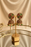 A pair of ornate gold-toned Indian jhumka earrings with green and red stone accents and dangling red and green beads, displayed on a gold stand on a glass table with a window and green foliage in the background.