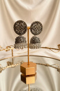 A pair of ornate silver-toned jhumka earrings with intricate beadwork and dangling bead strands, displayed on a gold stand on a reflective table with a window and greenery in the background.