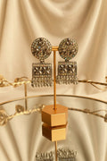 A pair of ornate silver-toned statement earrings displayed on a gold stand. The earrings feature circular studs with floral patterns set with iridescent stones, and rectangular dangle charms with dangling crystal fringe, shown on a reflective glass table with a window in the background.
