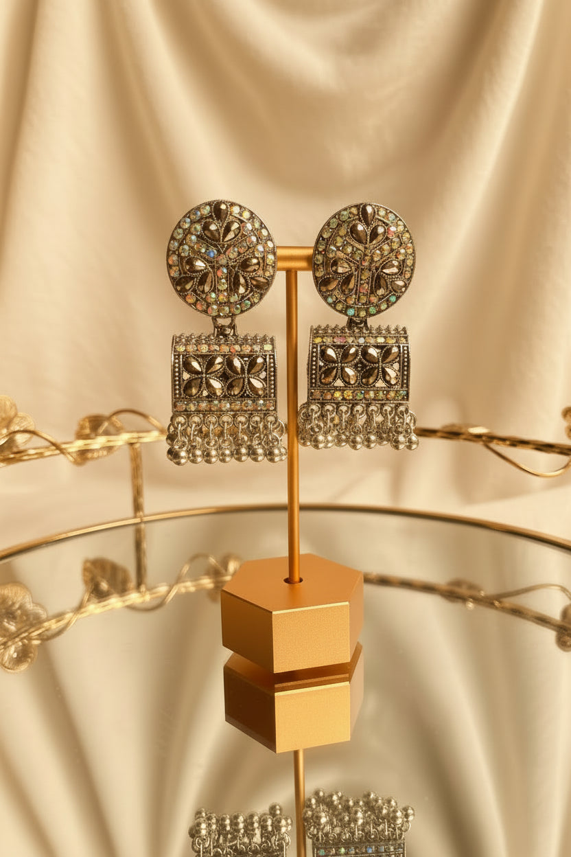 A pair of ornate silver-toned statement earrings displayed on a gold stand. The earrings feature circular studs with floral patterns set with iridescent stones, and rectangular dangle charms with dangling crystal fringe, shown on a reflective glass table with a window in the background.