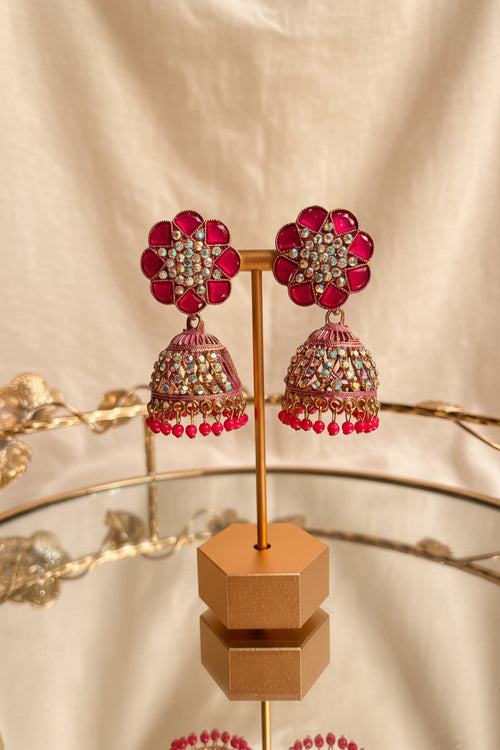 A pair of ornate Indian jhumka earrings displayed on a gold stand. The earrings feature bright pink floral studs with gold and white stones, hanging bell-shaped drops with pink stones, gold detailing, and dangling pink beads, set against a background of a window and greenery.