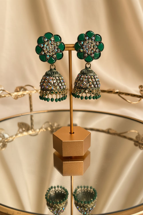A pair of ornate green and gold jhumka earrings displayed on a gold stand, set against a blurred background of a window and greenery.