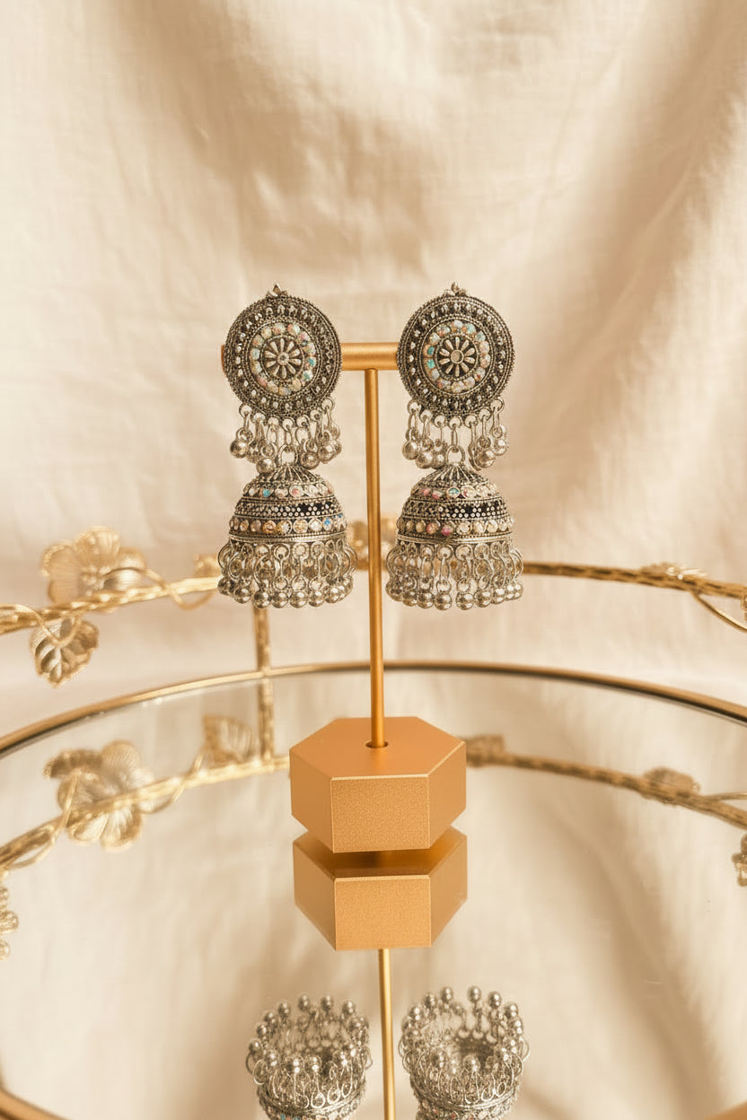 A pair of ornate silver-toned Indian jhumka earrings with intricate filigree work and colorful rhinestone accents, displayed on a gold stand on a glass table.