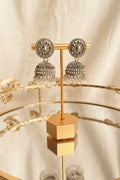 A pair of ornate silver-toned jhumka earrings displayed on a gold-colored stand, set against a softly blurred indoor background with warm wood tones and reflections.