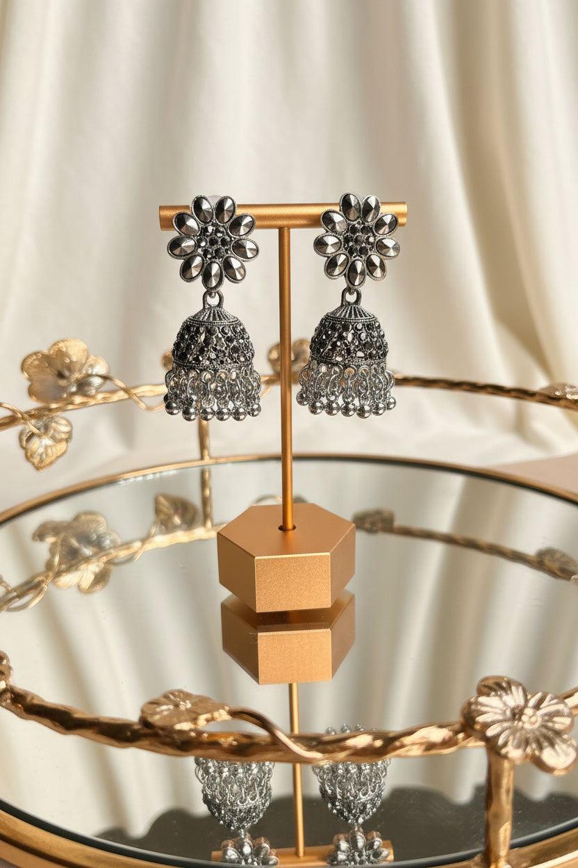 A pair of silver-toned Indian jhumka earrings displayed on a gold-colored stand. The earrings feature a floral stud design with small clear stones and a detailed bell-shaped dangle with intricate filigree work.
