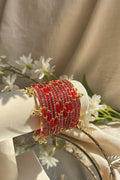 A stack of red and gold kashmiri bangles with red stones and gold-toned embellishments, displayed on a white jewelry stand in a bright indoor setting with a blurred vase and greenery in the background.