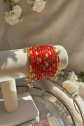 A stack of red kashmiri bangles with gold floral patterns and dangling gold charms, displayed on a cream jewelry stand in a sunlit indoor setting.