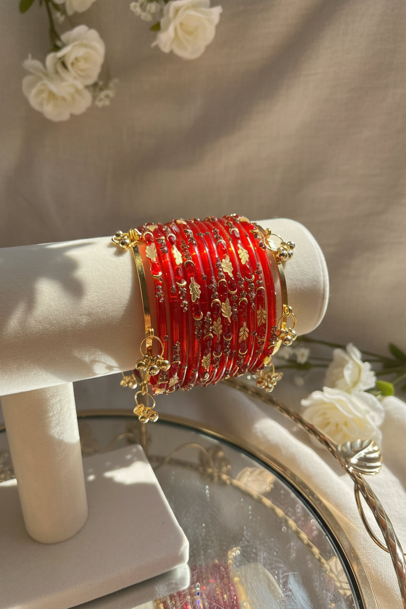 A stack of red kashmiri bangles with gold floral patterns and dangling gold charms, displayed on a cream jewelry stand in a sunlit indoor setting.