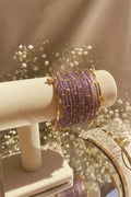 A set of wide lavender and gold beaded kashmiri bangles displayed on a cream jewelry stand, with a blurred indoor background featuring greenery and pink flowers.