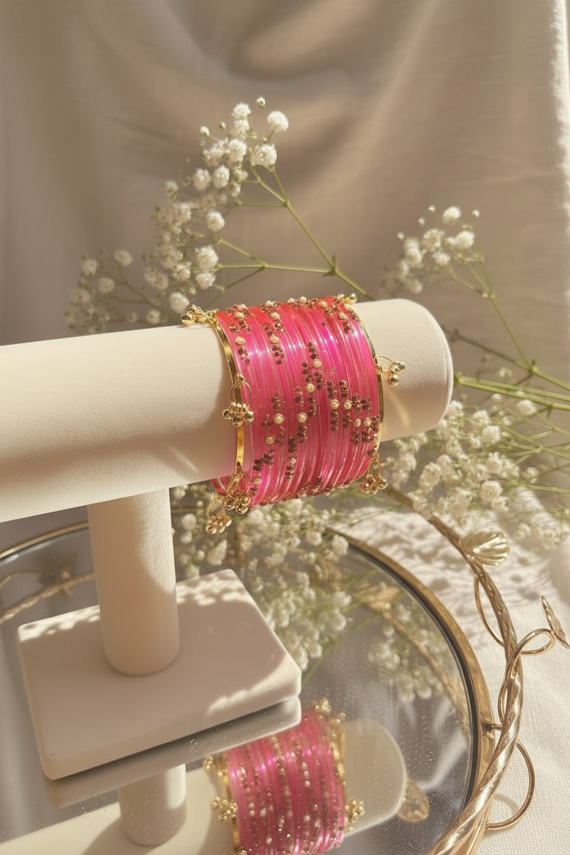 A stack of bright pink kashmiri bangles with gold-toned metal borders and small pearl-like bead accents, displayed on a cream jewelry stand in a softly lit indoor setting.