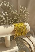 A stack of yellow beaded kashmiri bangles with gold-toned metal and small dark stones, displayed on a cream jewelry stand in a styled indoor setting with flowers and greenery in the background.