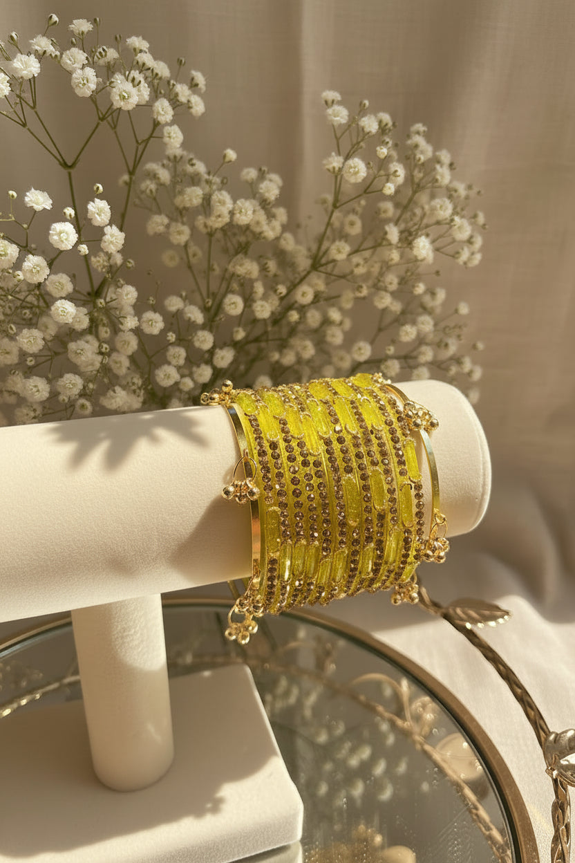 A stack of yellow beaded kashmiri bangles with gold-toned metal and small dark stones, displayed on a cream jewelry stand in a styled indoor setting with flowers and greenery in the background.