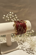 A stack of red kashmiri bangels with gold-toned metal borders and small pearl-like bead accents, displayed on a cream jewelry stand in a bright indoor setting with blurred greenery and pink flowers in the background.