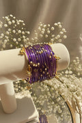 A set of purple glass kashmiri bangles with gold-plated borders, small gold beads, and white pearl-like accents, displayed on a cream-colored jewelry stand.