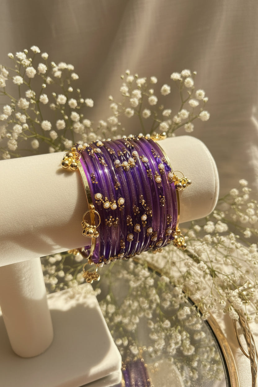 A set of purple glass kashmiri bangles with gold-plated borders, small gold beads, and white pearl-like accents, displayed on a cream-colored jewelry stand.