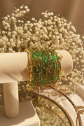 A set of gold-plated kashmiri bangles adorned with multiple rows of green emerald-cut crystals, displayed on a cream-colored jewelry stand.