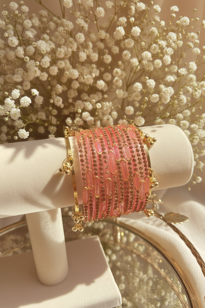 A stack of pink beadedkashmiri bangles with gold-toned metal borders and small dangling gold charms, displayed on a cream jewelry stand in a softly lit indoor setting with blurred greenery and flowers in the background.