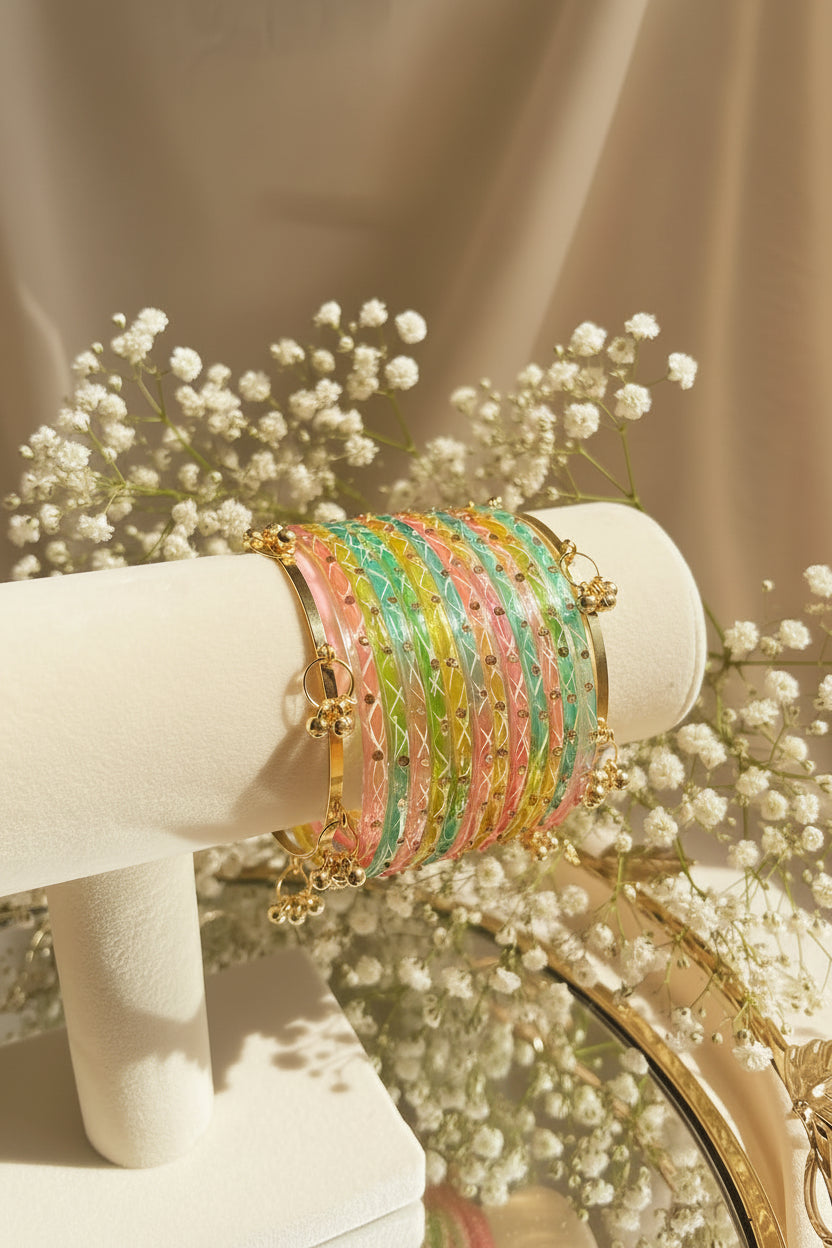 A set of pastel-colored woven kashmiri bangles with gold-toned metal details, displayed on a cream jewelry stand in a styled indoor setting with flowers and greenery in the background.