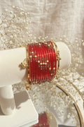 A set of red kashmiri bangles with gold-toned borders and beadwork, displayed on a cream-colored jewelry stand.