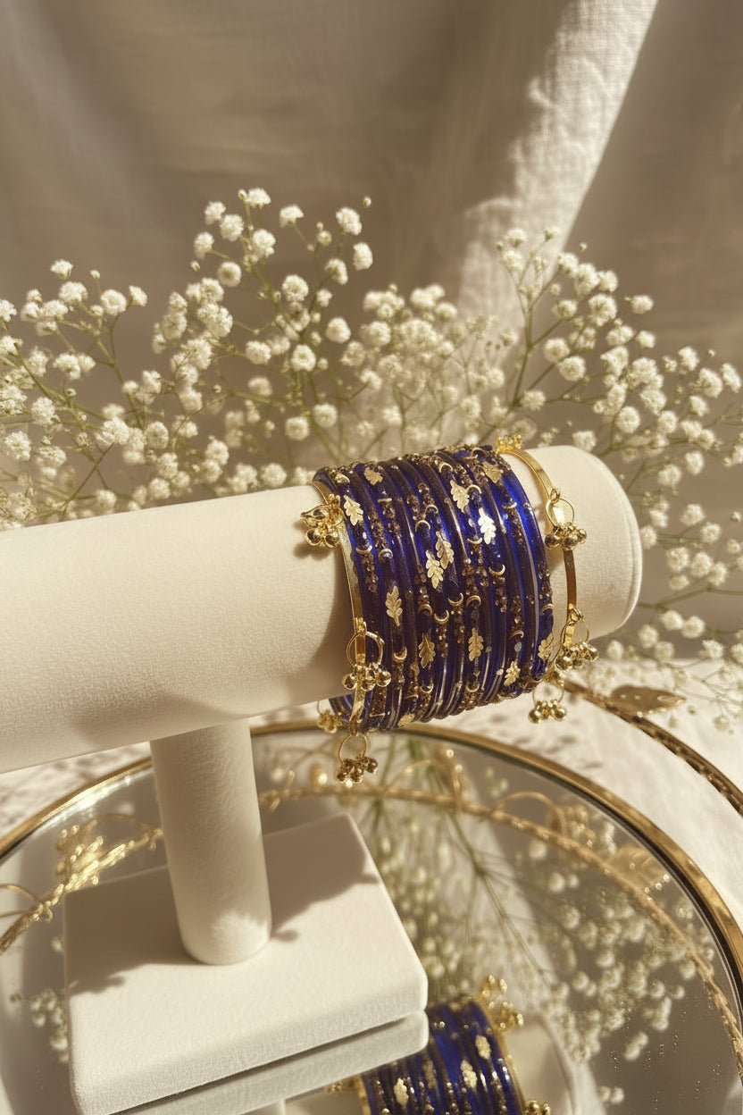 A stack of royal blue beaded kashmiri bangles with gold foil patterns and gold-toned chain charms, displayed on a cream-colored jewelry stand.