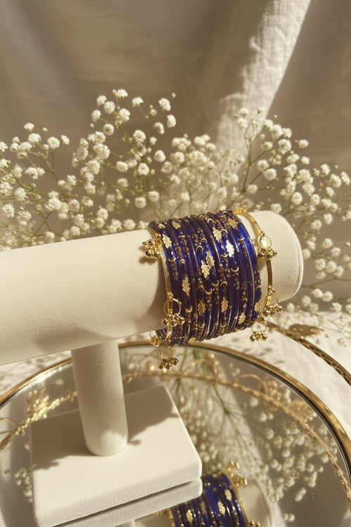 A stack of royal blue beaded kashmiri bangles with gold foil patterns and gold-toned chain charms, displayed on a cream-colored jewelry stand.