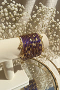 A stack of purple and gold beaded kashmiri bangles displayed on a cream-colored jewelry stand, with a blurred background showing green foliage and pink flowers.