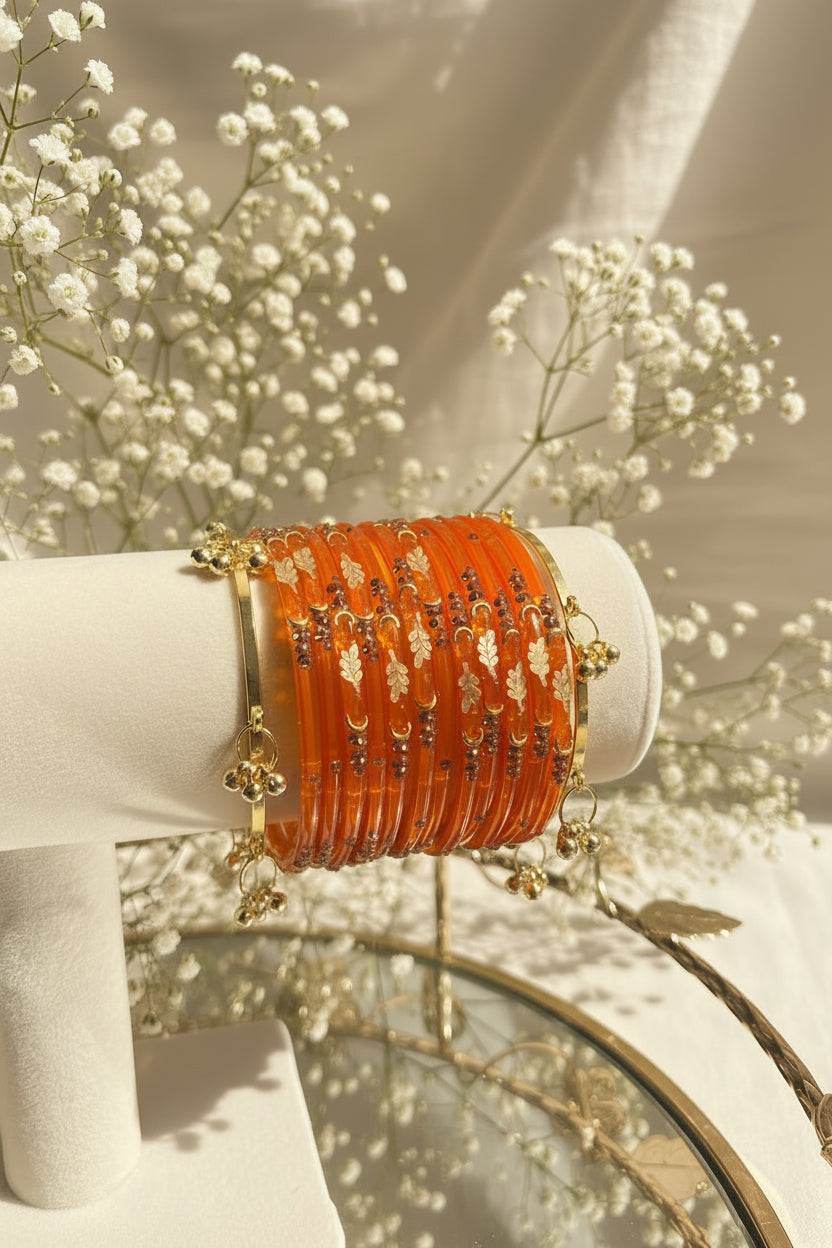 A stack of orange kashmiri bangles with gold and silver decorative elements displayed on a white jewelry stand, with a blurred background of flowers and greenery.