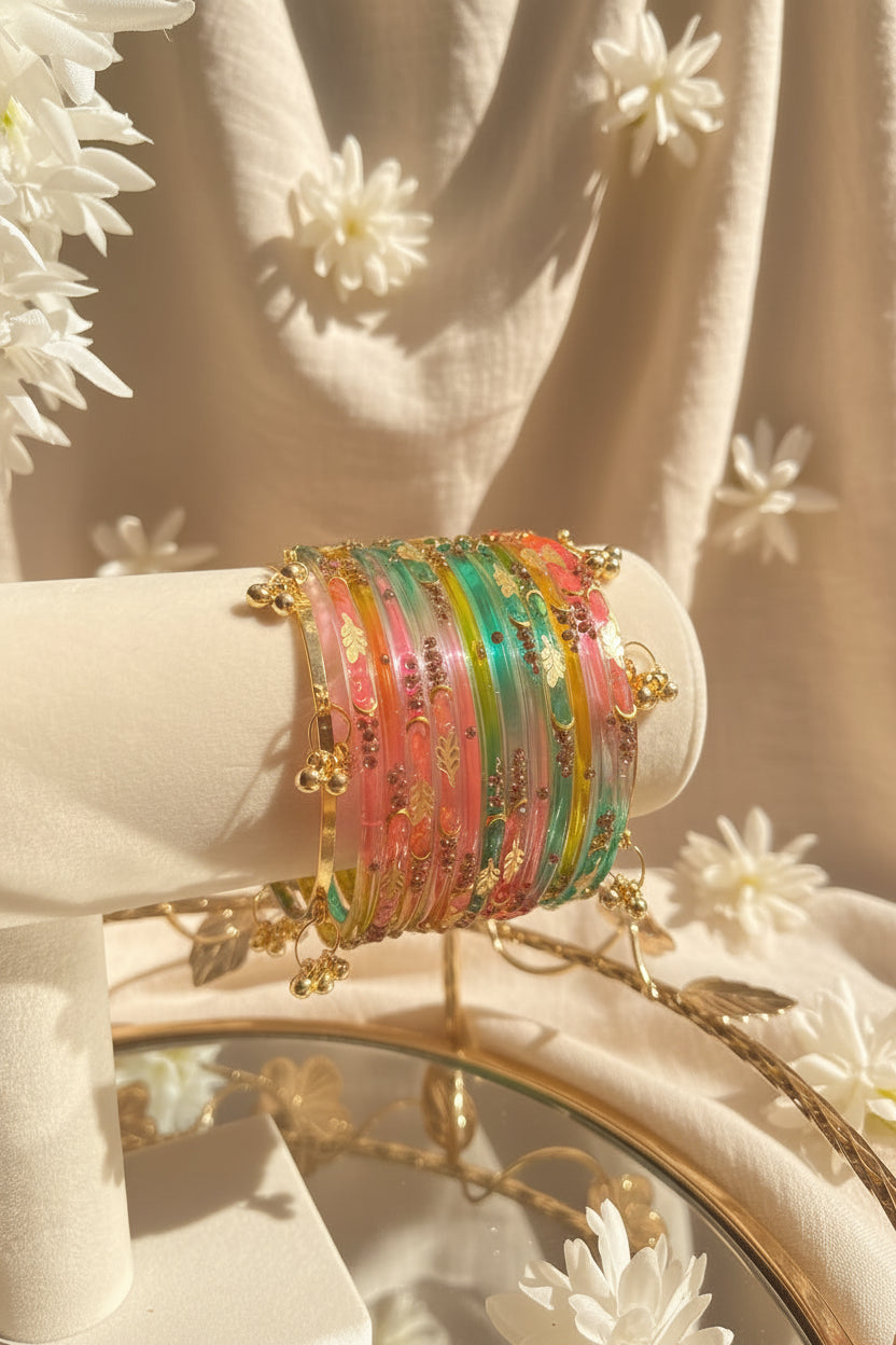 A set of colorful kashmiri bangles with gold-toned beads and charms displayed on a cream-colored jewelry stand. The background features a blurred glass vase with green leaves and pink flowers in a sunlit setting.