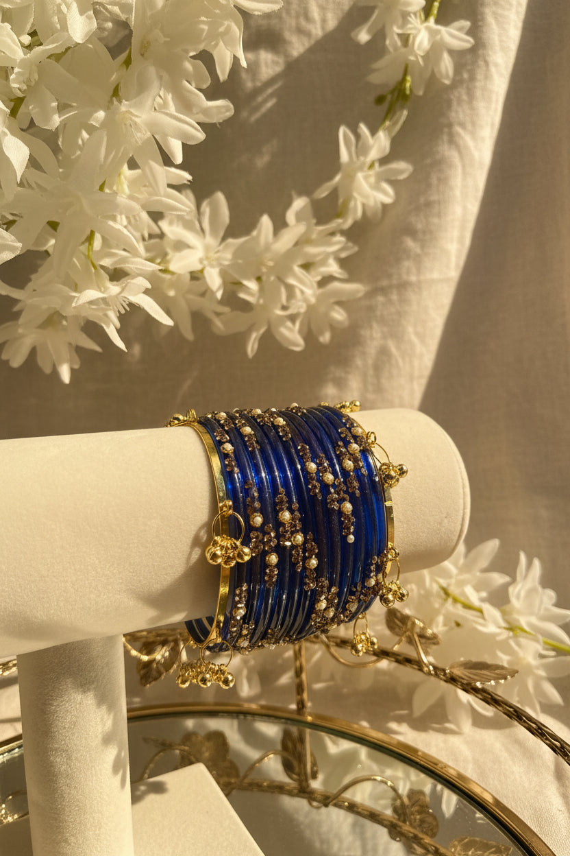 A set of royal blue beaded kashmiri bangles with gold-plated borders, pearls, and small gold floral charms, displayed on a cream-colored velvet stand.