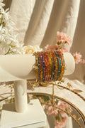 A stack of colorful kashmiri bangles with gold-toned metal and dangling charms, displayed on a cream-colored jewelry stand. The background features a blurred glass vase with green leaves and white flowers.