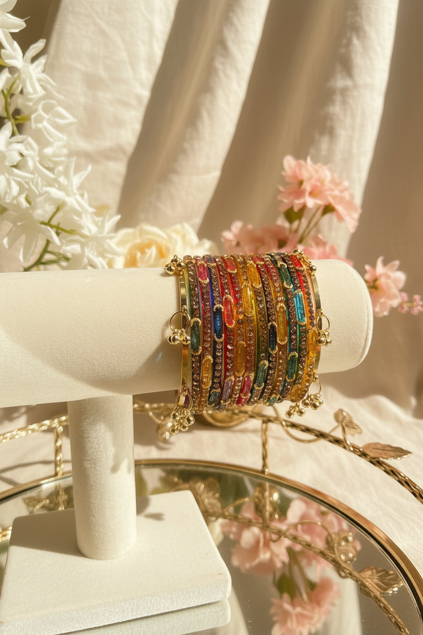 A stack of colorful kashmiri bangles with gold-toned metal and dangling charms, displayed on a cream-colored jewelry stand. The background features a blurred glass vase with green leaves and white flowers.