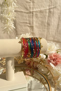 A stack of colorful kashmiri bangles with gold-toned hardware and bead details displayed on a cream-colored jewelry stand. The background features a blurred window view with green foliage and pink flowers.