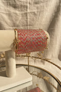 A pink and silver beaded kashmiri bangels with gold-toned hardware displayed on a white jewelry stand, with flowers and a mirror in the background.