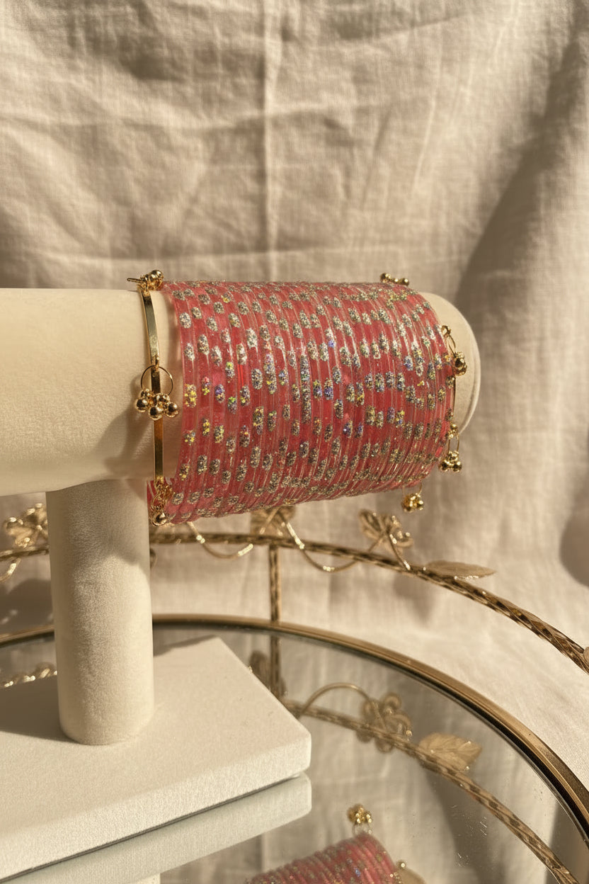 A pink and silver beaded kashmiri bangels with gold-toned hardware displayed on a white jewelry stand, with flowers and a mirror in the background.