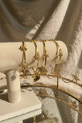 A set of gold-toned ghungroo bell bangle bracelets displayed on a cream-colored jewelry stand, with a glass vase of flowers in the background.
