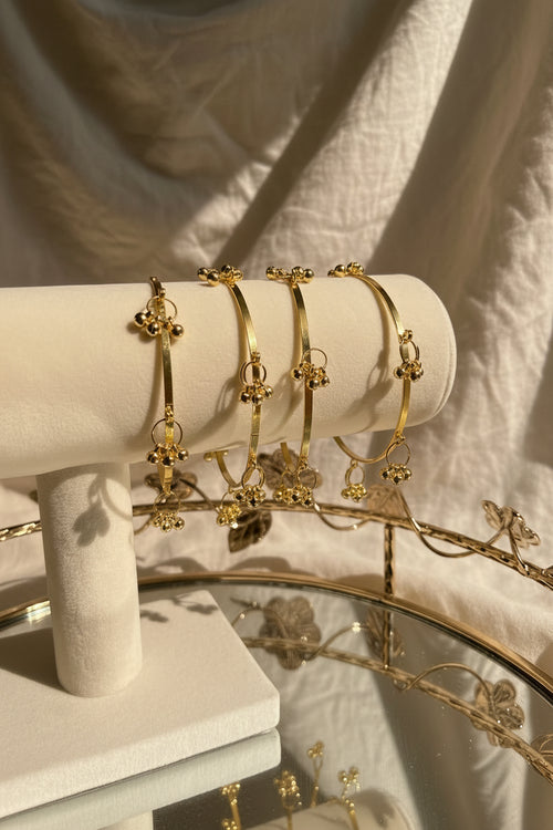 A set of gold-toned ghungroo bell bangle bracelets displayed on a cream-colored jewelry stand, with a glass vase of flowers in the background.